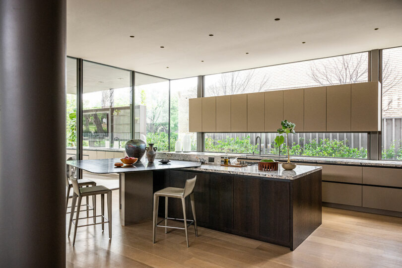 Modern kitchen with large island, bar stools, light wood floors, minimalist cabinetry, and large windows overlooking a garden.