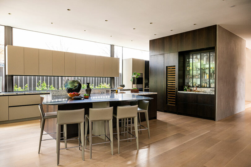 Modern kitchen with light wood floors, beige cabinets, a large island with four chairs, and floor-to-ceiling windows letting in natural light.
