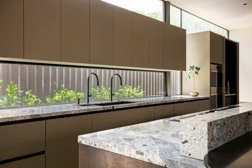 Modern kitchen with terrazzo countertops, minimalist cabinetry, two sinks with tall faucets, and a long horizontal window overlooking greenery.
