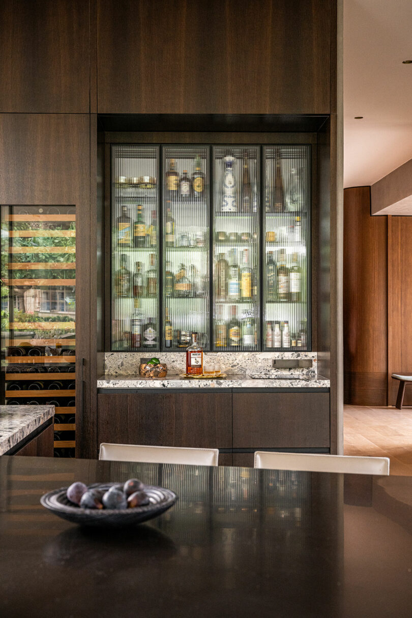 A modern home bar with glass-front cabinets displaying various bottles, a marble countertop, and a bowl of fruit on a dark dining table in the foreground.