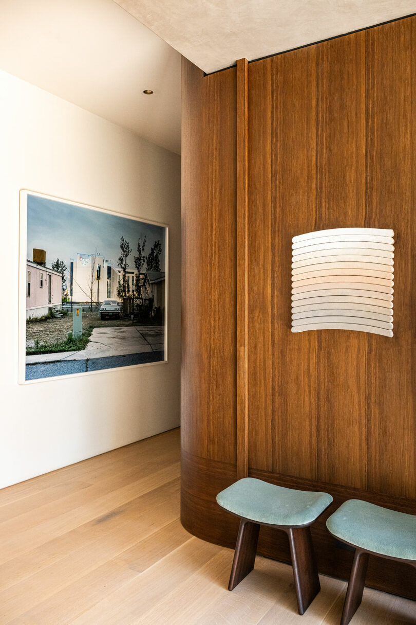 A modern interior with wood-paneled walls, two blue cushioned stools, a wall-mounted light fixture, and a large framed photograph of houses on the opposite wall.