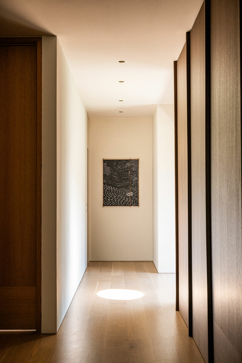 A modern hallway with light wood floors, white walls, recessed ceiling lights, and a framed artwork at the end, illuminated by natural sunlight.