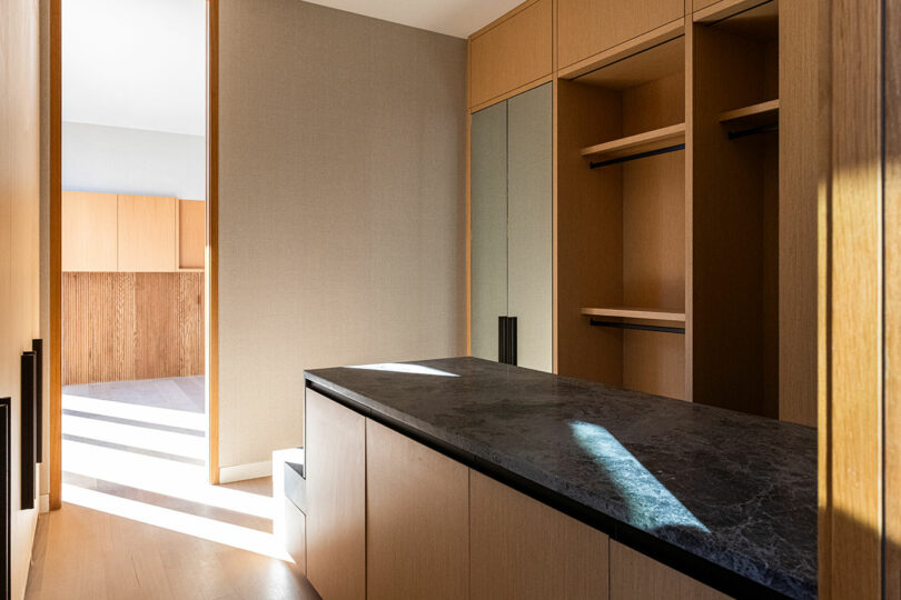 A modern walk-in closet with wooden shelves, a dark stone countertop, and natural sunlight streaming in through a doorway.