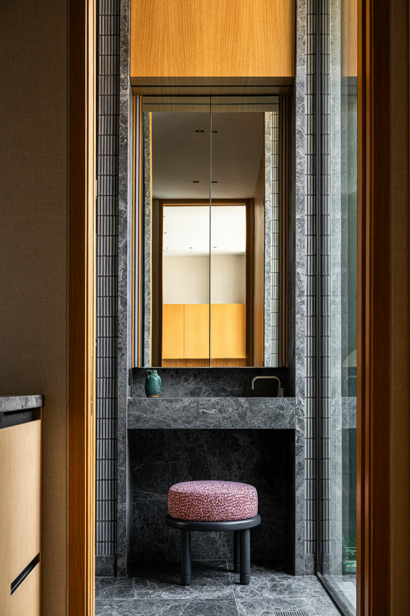 A modern bathroom with a marble vanity, large mirror, small red patterned stool, and a narrow window letting in natural light.