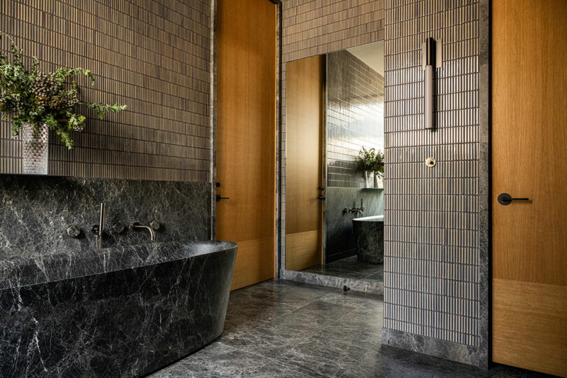 Modern bathroom with dark marble bathtub, matching vanity, vertical wall tiles, large mirror, wooden doors, and a wall-mounted sconce.