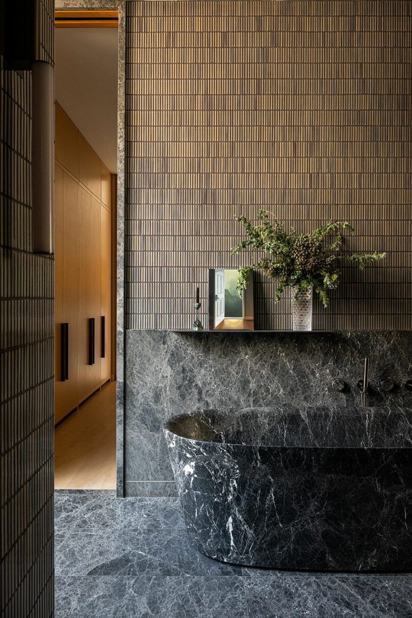 A modern bathroom with dark gray marble tub and floor, vertical tile walls, a vase with green branches, and an open doorway to a light wood hallway.
