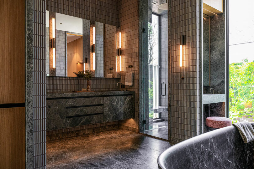 Modern bathroom with dark marble flooring, dual sinks, large mirrors, vertical wall sconces, glass shower, and a freestanding bathtub next to a window with greenery outside.