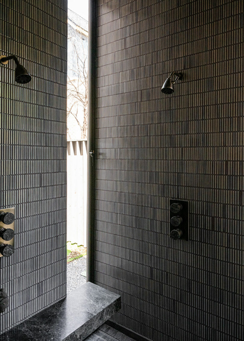 Modern shower with vertical gray tile walls, two showerheads, control knobs, and a built-in bench; a narrow window provides natural light and an outdoor view.