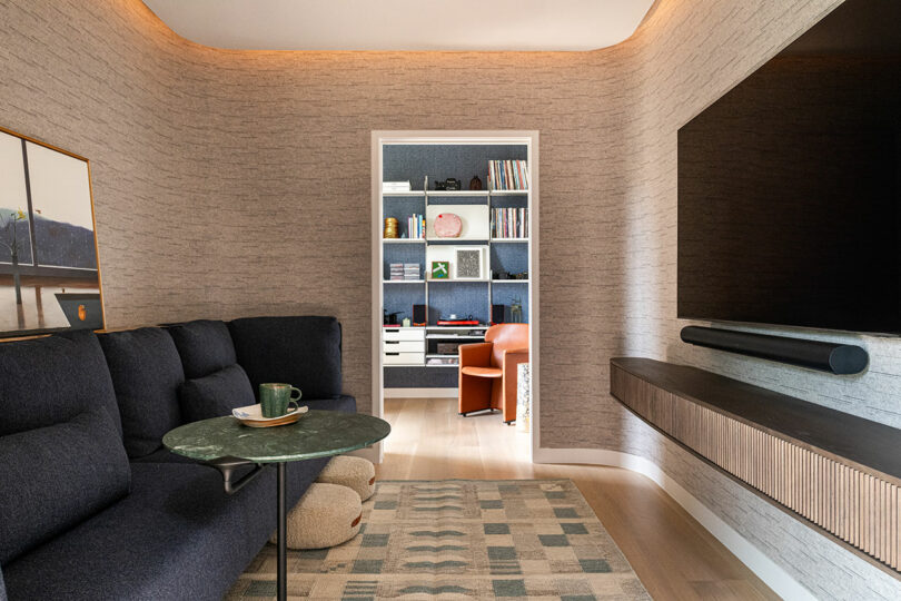 Modern living room with a dark sectional sofa, green marble coffee table, wall-mounted TV, and a view into a home office with shelves, desk, and an orange chair.