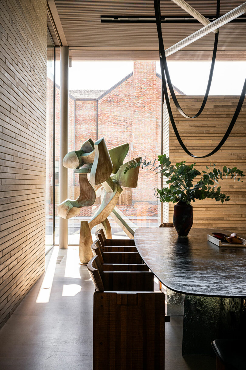 Modern dining area with wooden chairs, a dark table topped with a vase of greenery, large abstract sculpture, and floor-to-ceiling windows showing a brick building outside.