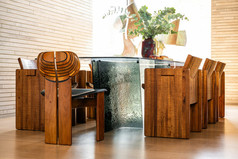 A glass dining table with a textured base is surrounded by eight wooden chairs with curved backs; a vase with greenery sits on the table.