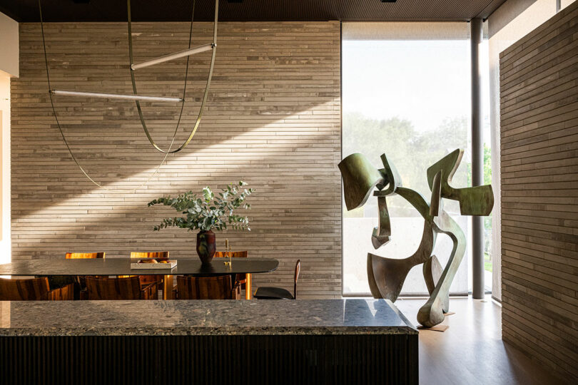 Modern dining area with a large stone table, wooden chairs, a minimalist light fixture, a plant centerpiece, and an abstract metal sculpture near a tall window.