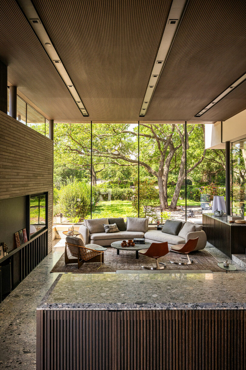 Modern living room with large floor-to-ceiling windows, a round sofa, two brown chairs, and views of a lush green garden outside. Natural light fills the space.