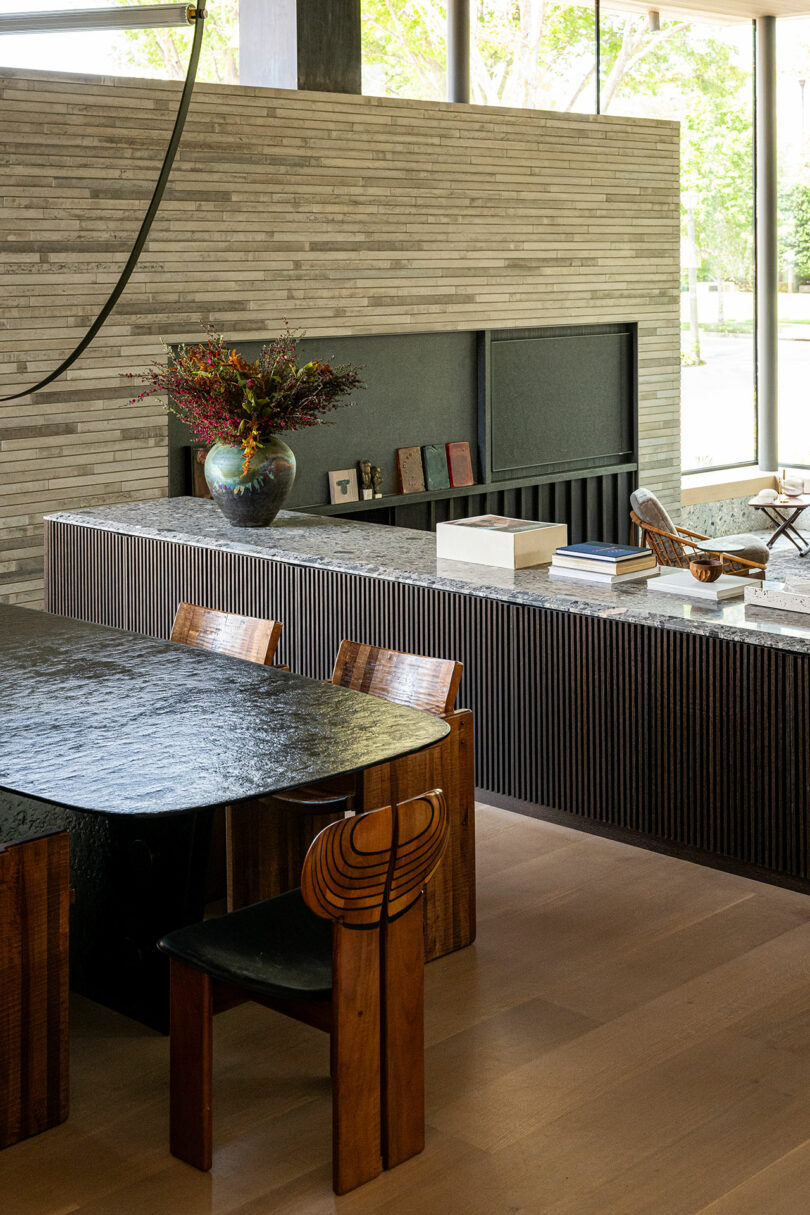 Modern dining area with a black textured table, wooden chairs, a countertop with a vase of flowers, books, and decorative items, set against a neutral-toned wall with large windows.