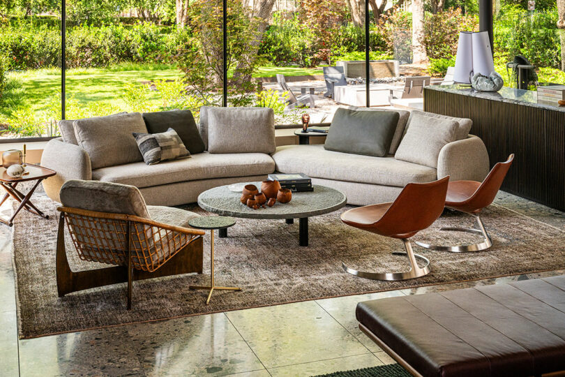 Modern living room with a curved beige sofa, two brown chairs, a round coffee table, and floor-to-ceiling windows overlooking a green garden.