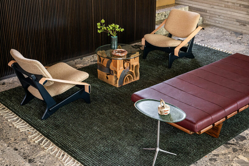 A modern living room with two beige armchairs, a round glass coffee table, a maroon leather bench, a small side table, and a dark textured rug on a stone floor.