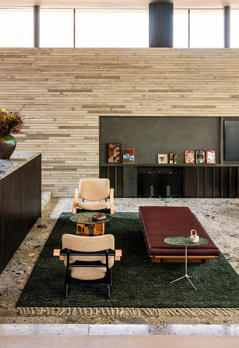 Modern living room with two armchairs, a maroon daybed, a small side table, and decorative art on a stone shelf against a textured wall.