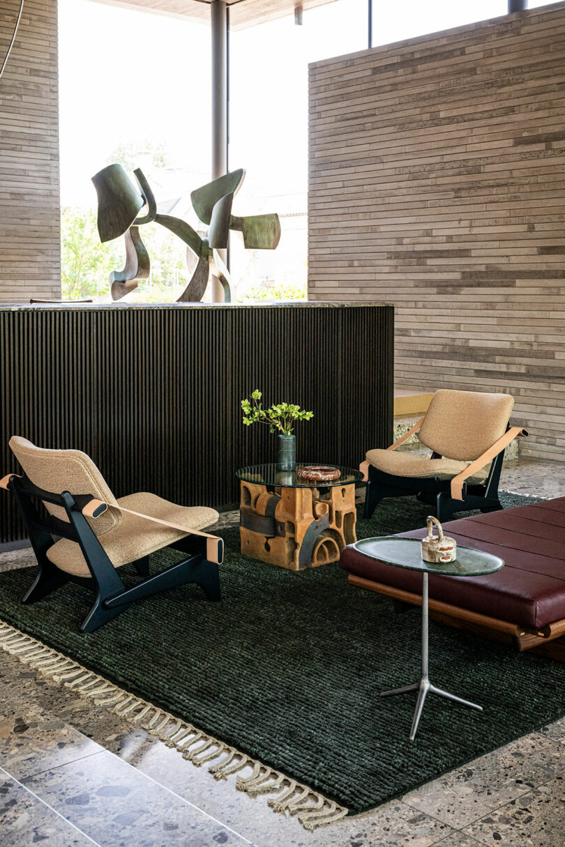 Modern lounge area with two armchairs, a leather bench, a round side table, a sculptural wooden coffee table, and abstract art, set on a dark rug over a stone floor.