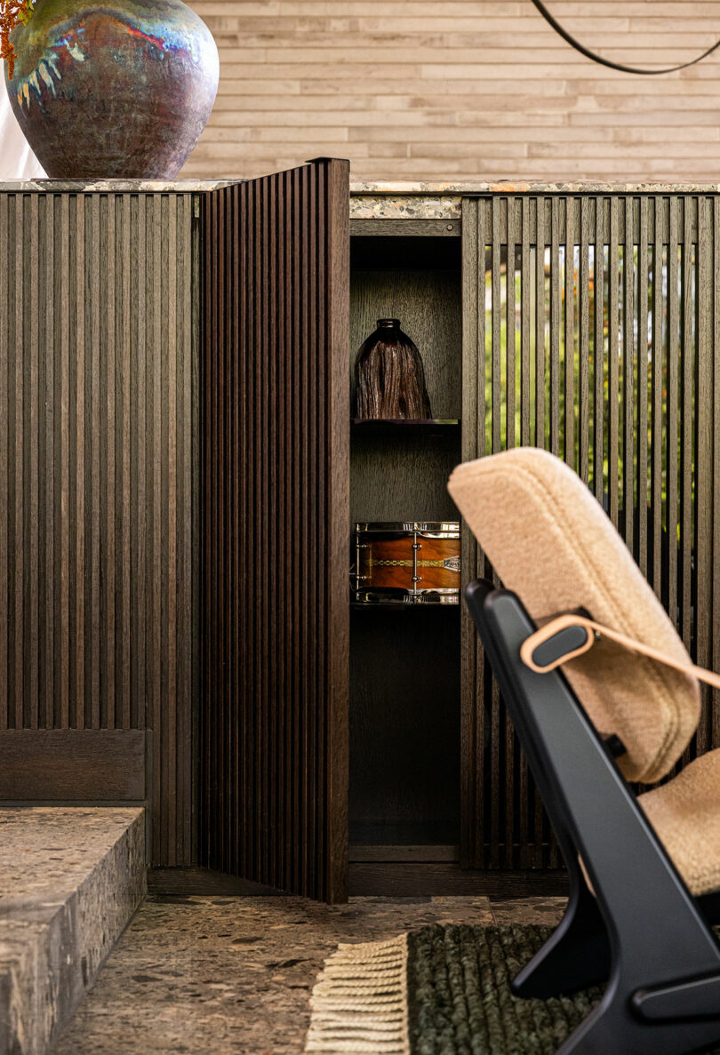 A wooden slatted cabinet with an open door reveals two shelves holding a dark vase and a decorative box; a beige chair and a large ceramic pot are in the foreground.