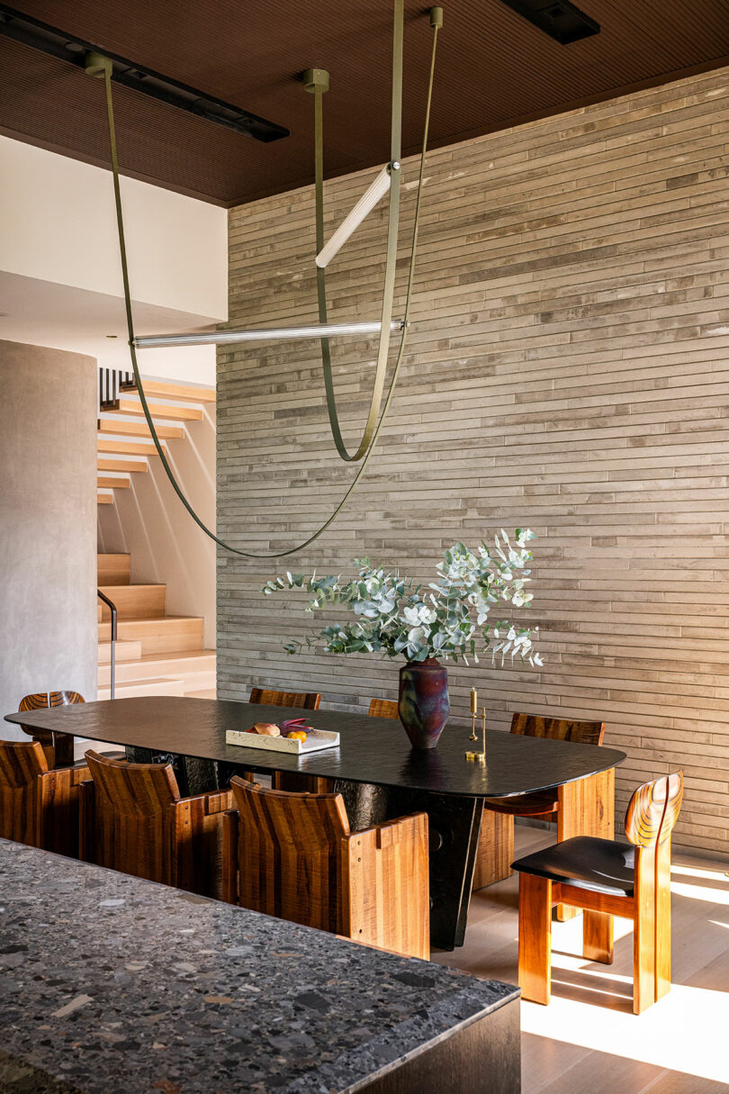 A modern dining room with a dark wooden table, matching chairs, a vase with greenery, books, and a textured stone wall in the background.
