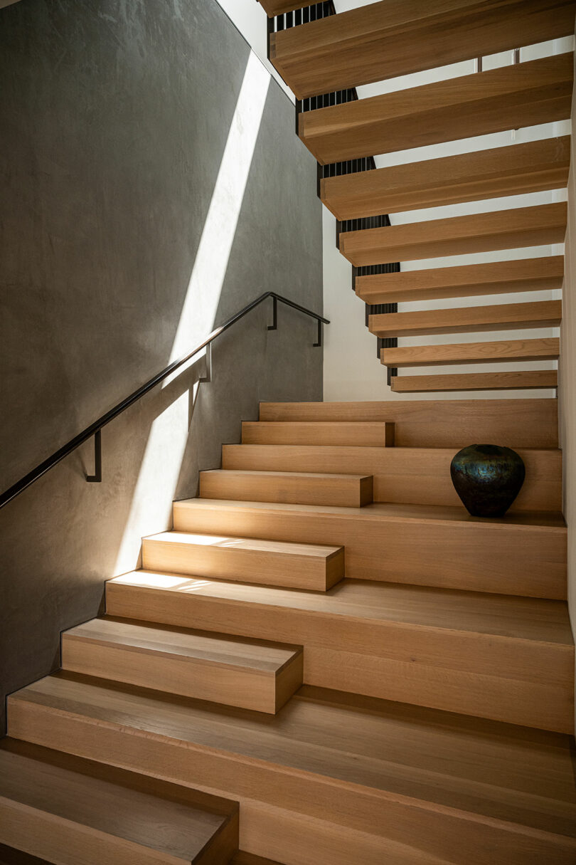 Wooden floating staircase with wide steps, a simple black handrail, a gray textured wall, and a ceramic vase placed on one of the lower steps. Sunlight streams in from above.