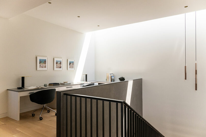 A modern home office with a white desk, black chair, three framed pictures on the wall, and sunlight streaming in from above a staircase with black railing.