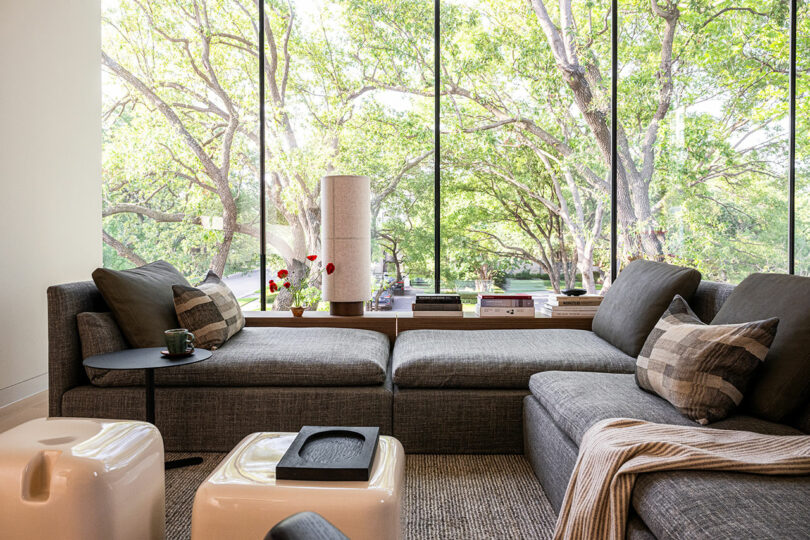 A modern living room with gray sectional sofa, plaid pillows, a floor lamp, and large windows overlooking green trees outside.