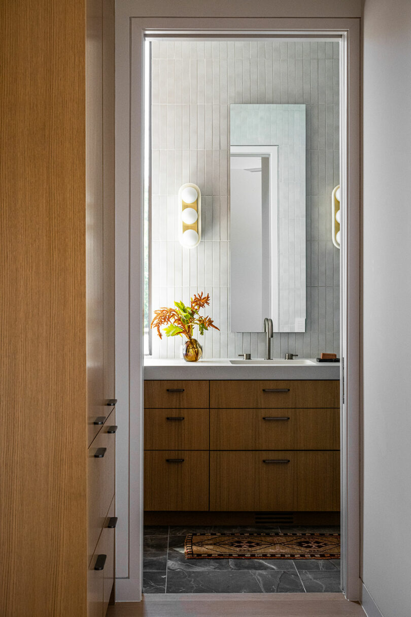 A modern bathroom with wooden cabinets, a rectangular mirror, wall sconces, a small vase of orange flowers, and a patterned rug on a dark tile floor.