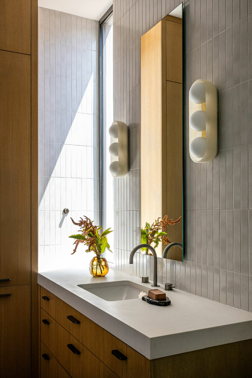 Modern bathroom with a white countertop, built-in sink, wooden cabinets, vertical mirror, wall light fixtures, and a glass vase with foliage near a tall window.