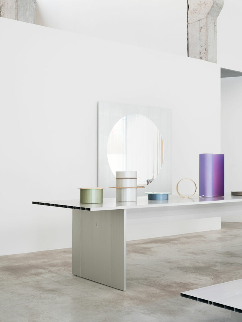 A minimalist room with a long white table displaying various cylindrical and geometric objects in pastel and metallic colors; a circular mirror hangs on the wall behind.