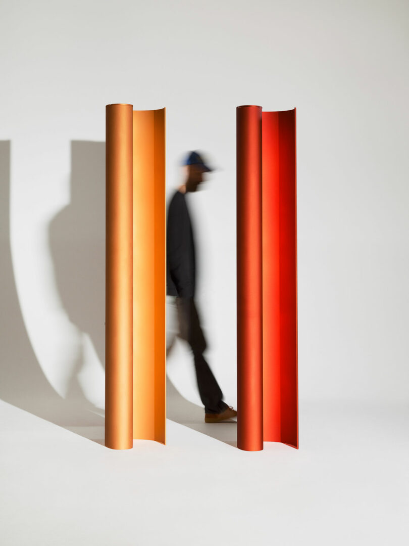 A person walks past two large, upright, curved panels in orange and red tones against a plain background.