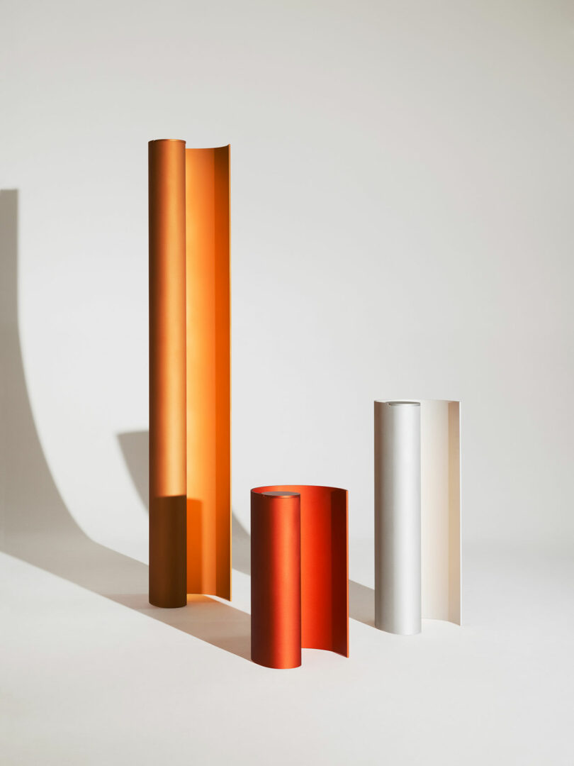 Three upright, curved sheets of colored material—orange, red, and white—are arranged on a plain surface, casting soft shadows against a light background.
