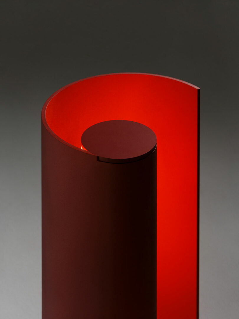 A modern, cylindrical red lamp with an open spiral design emits a warm red light, set against a neutral gray background.