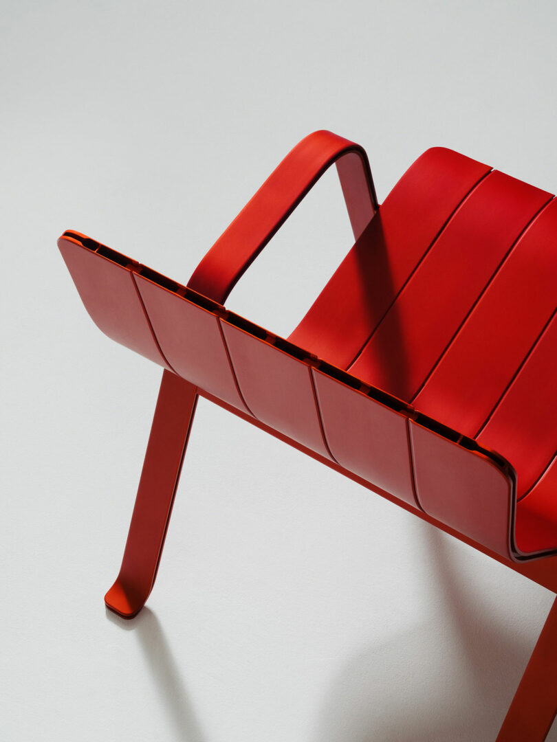 A modern red metal chair with a slatted seat, angled armrests, and slightly curved legs, photographed against a plain white background.