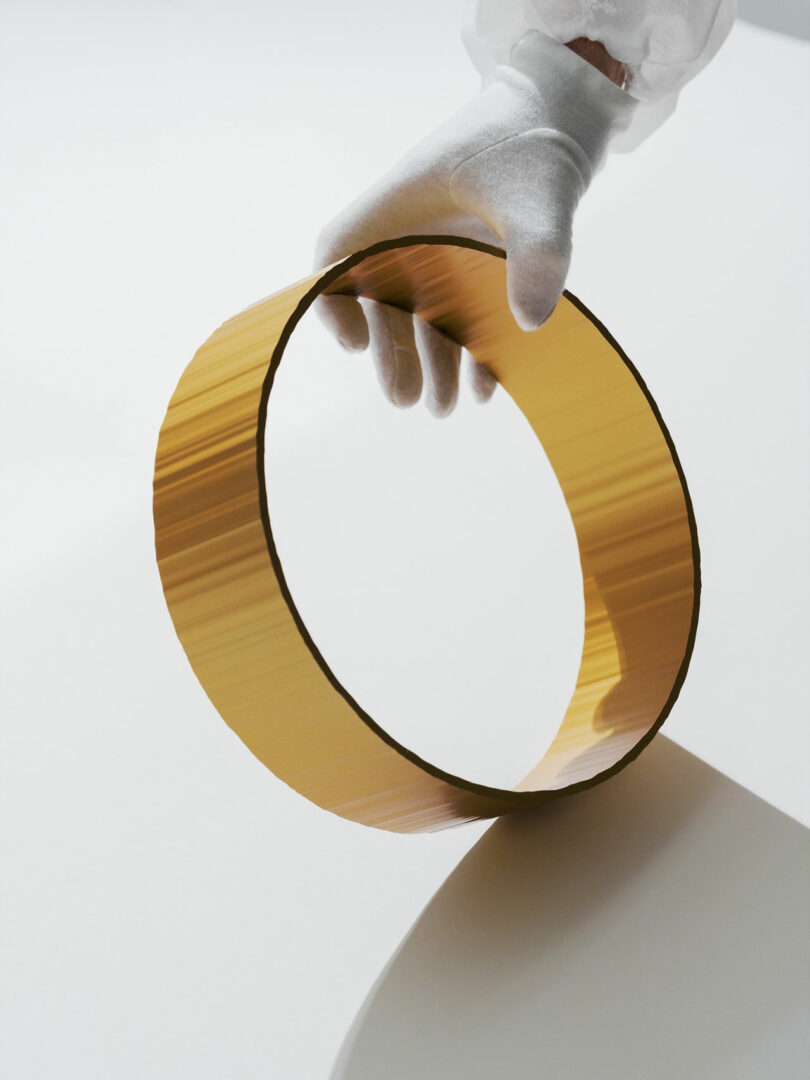 A gloved hand holds a large, thin, circular, gold-colored wafer upright, casting a shadow on a white surface.