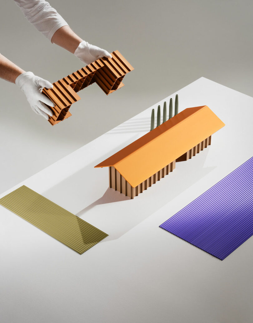 A person wearing white gloves places a striped, brown architectural model next to a similar structure with an orange roof, on a white surface with colored panels.