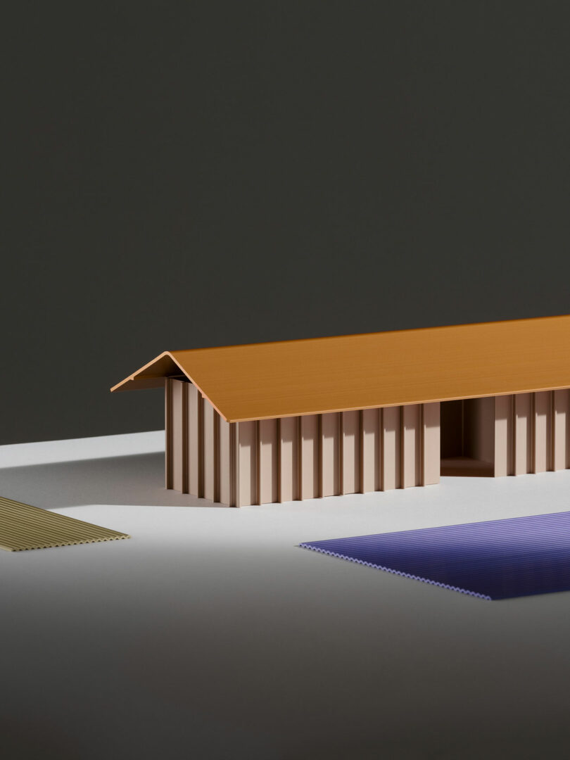 A minimalist model of a rectangular building with a brown slanted roof, beige vertical walls, and a cut-out doorway, placed on a plain surface with colored mats nearby.