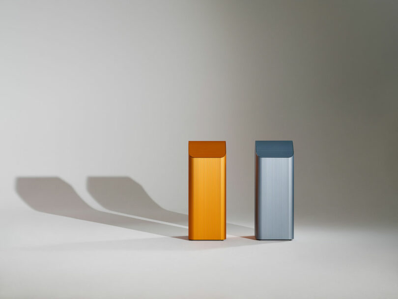 Two rectangular, vertically ribbed bins—one orange and one gray—stand side by side on a plain surface, casting long shadows to the left.