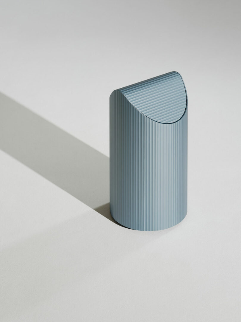 A cylindrical blue object with vertical ridges and a curved top sits on a light surface, casting a long shadow to the left.