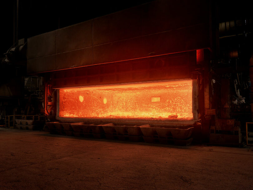 A large industrial furnace glows with intense orange heat in a dimly lit factory setting, with metal trays positioned beneath its opening.