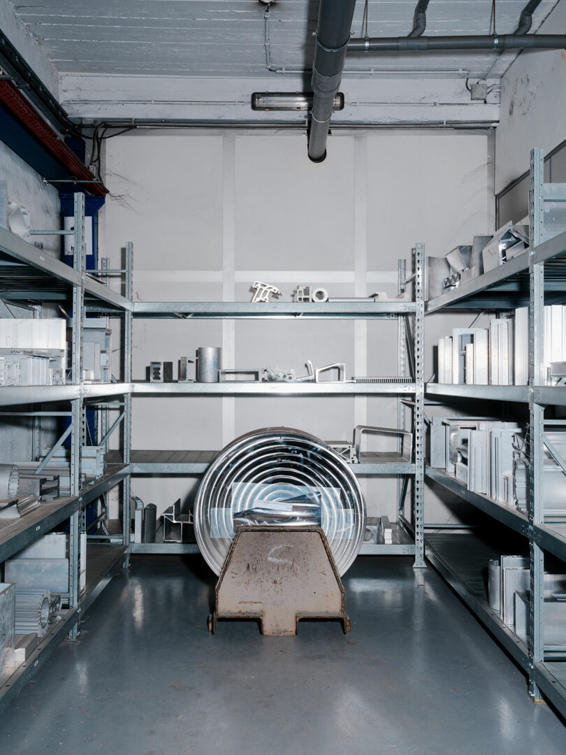 Industrial storage room with metal shelves holding various metal parts and ductwork; large coiled metal pipe and a container positioned in the center foreground.