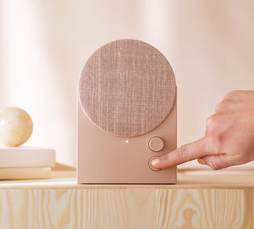 A hand presses a button on a modern, beige-colored speaker with a fabric-covered circular front, placed on a light wooden surface