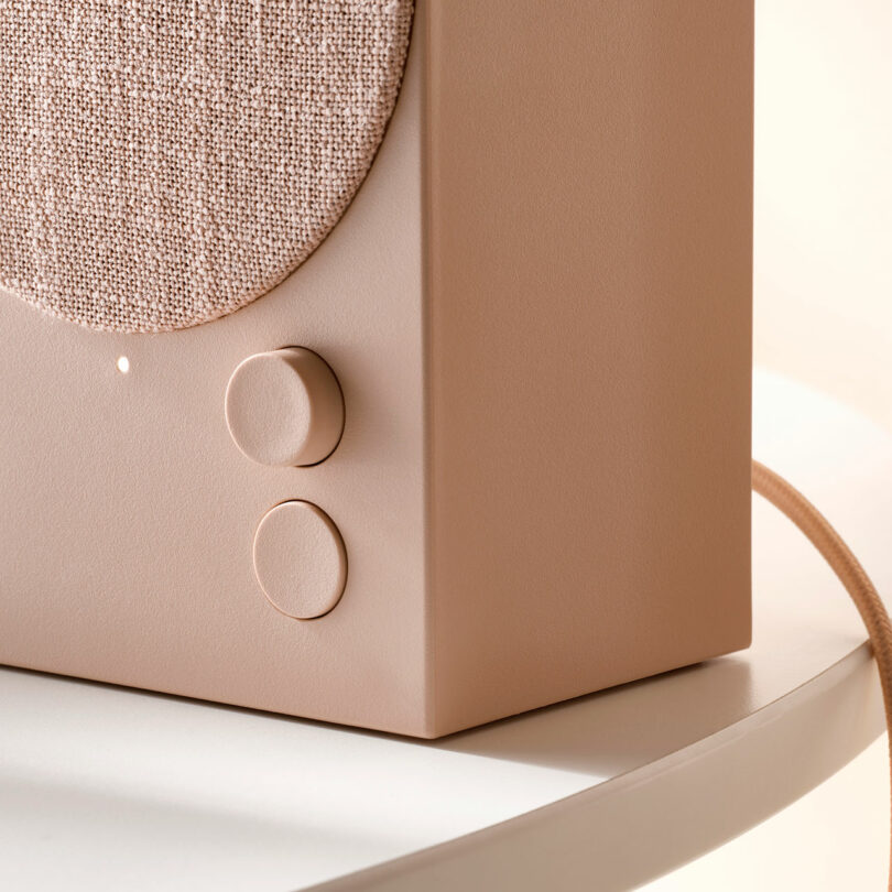 Close-up of a beige speaker with a fabric grille, two round control knobs, and a fabric-covered power cord on a white surface