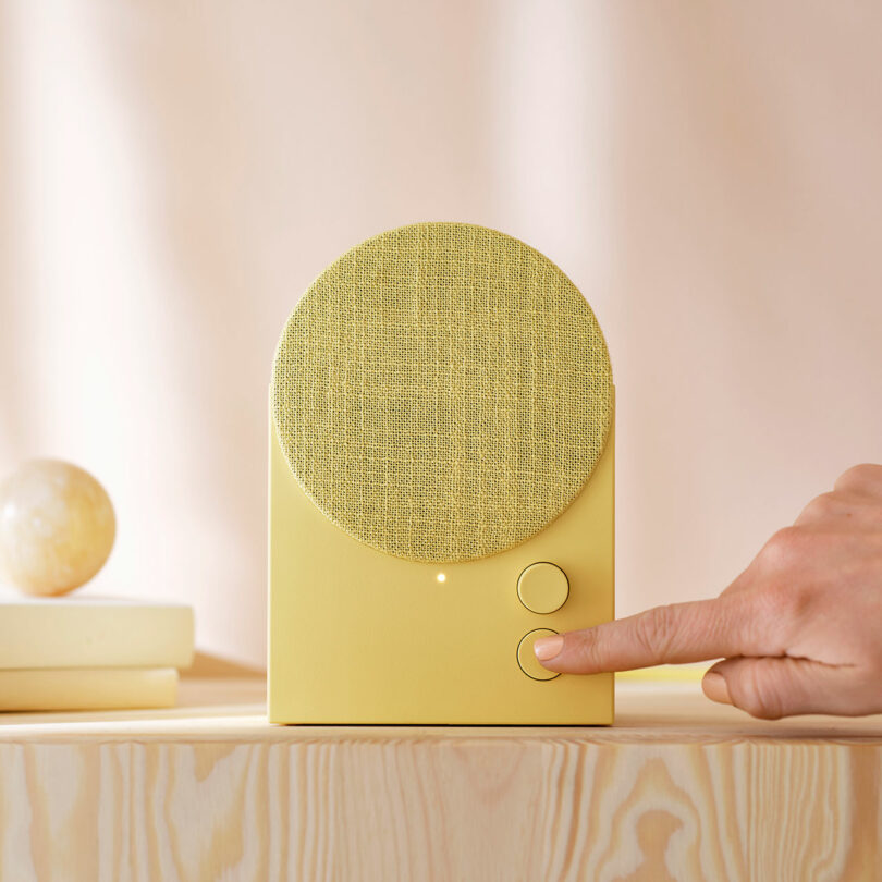 A hand presses a button on a yellow tabletop speaker with a fabric-covered circular front, placed on a light wooden surface