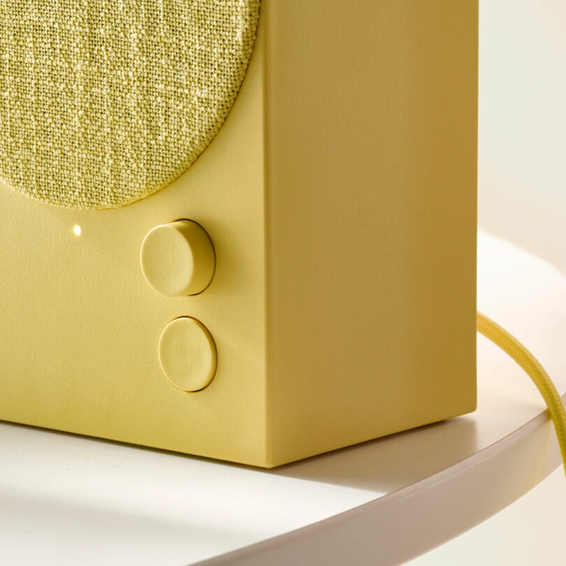 A close-up of a yellow speaker with fabric grille, two round control knobs, and a yellow cord, placed on a white surface