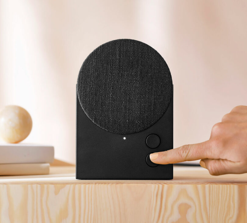 A hand presses a button on a modern black speaker with a circular fabric cover, placed on a light wooden surface
