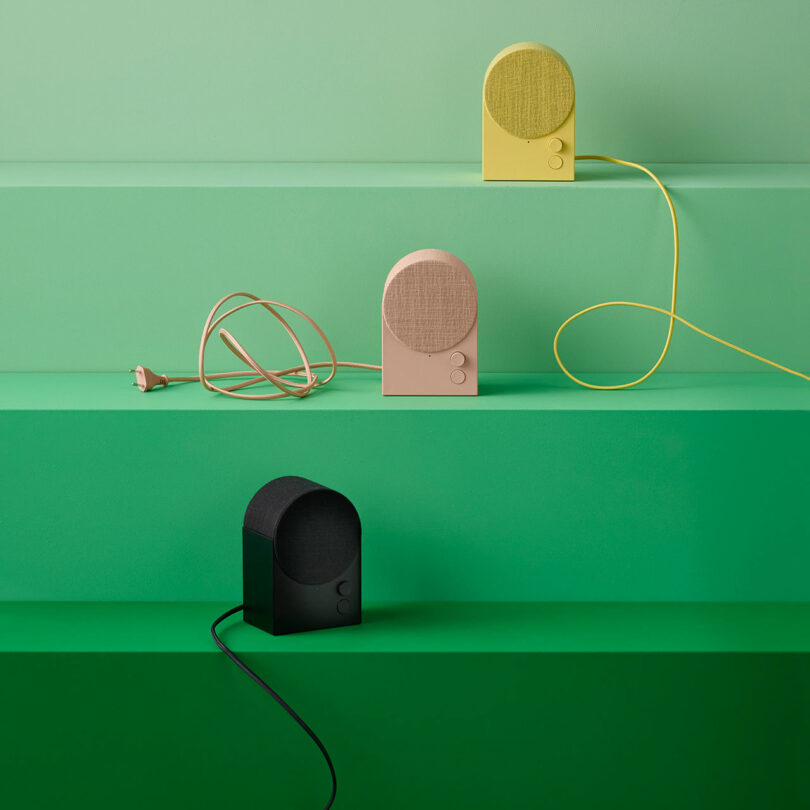 Three small tabletop speakers in yellow, beige, and black sit on green shelves with their power cords visible against a minimalist green background