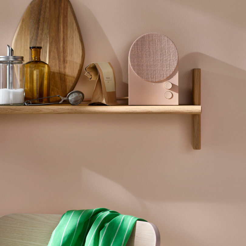 A wooden shelf with kitchen items, including a jar, brown bottle, cutting boards, paper bag, and a modern beige speaker, above a chair draped with a green-striped cloth