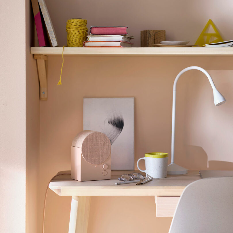 A small desk with a lamp, mug, eyeglasses, and a speaker sits below a shelf holding books, string, and geometric decor against a light-colored wall