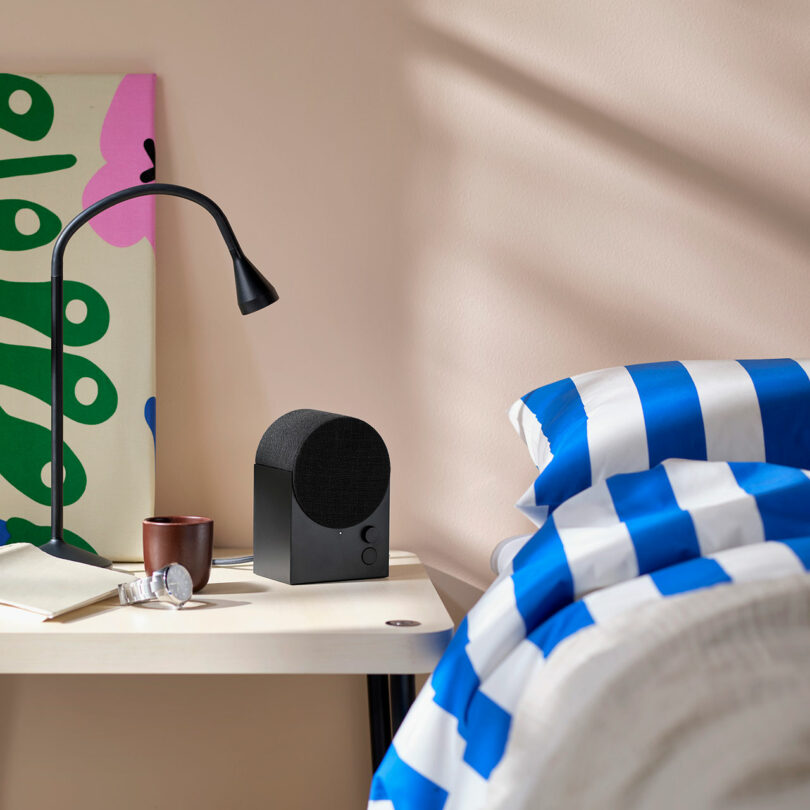 A modern bedside table with a black lamp, a speaker, a brown mug, and a pair of glasses next to a bed with blue and white stripe bedding